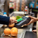 young adult man holding plastic credit card in hand, using terminal and paying for shopping in supermarket