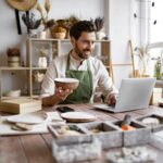 A small business owner sits at a desk in his place of business holding a bowl with one hand and using his laptop with his other hand