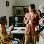 Group of three diverse gen Z colleagues wearing stylish outfits chatting during coffee break in office