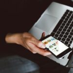 A person holds a smart phone in their hand and a laptop on their lap.