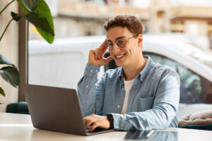 young man with curly hair and glasses smiling at his laptop in a bright room with a plant