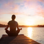 woman meditating on wooden pier near lake at sunset