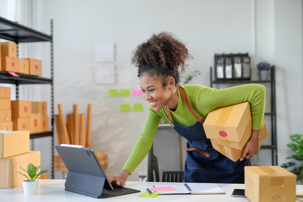 Young entrepreneur is managing her small business from her home office, smiling while carrying packages and using a digital tablet