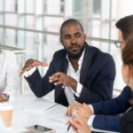 A man sitting at a table surrounded by coworkers, explaining something.