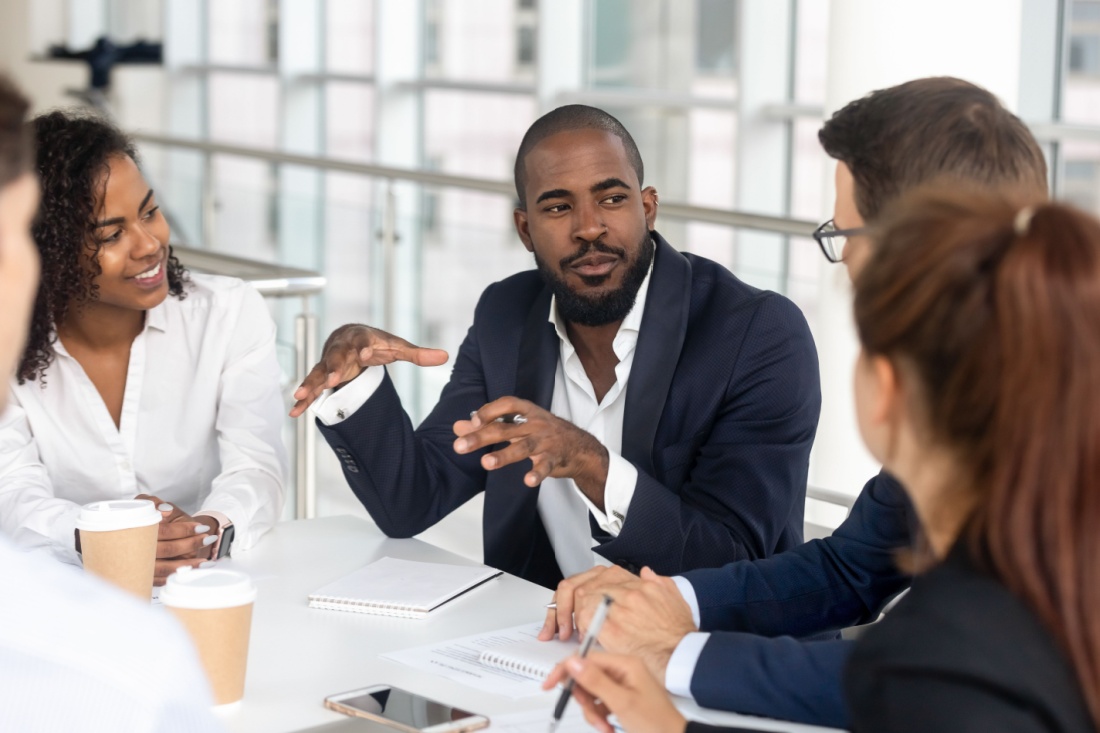 A man sitting at a table surrounded by coworkers, explaining something.