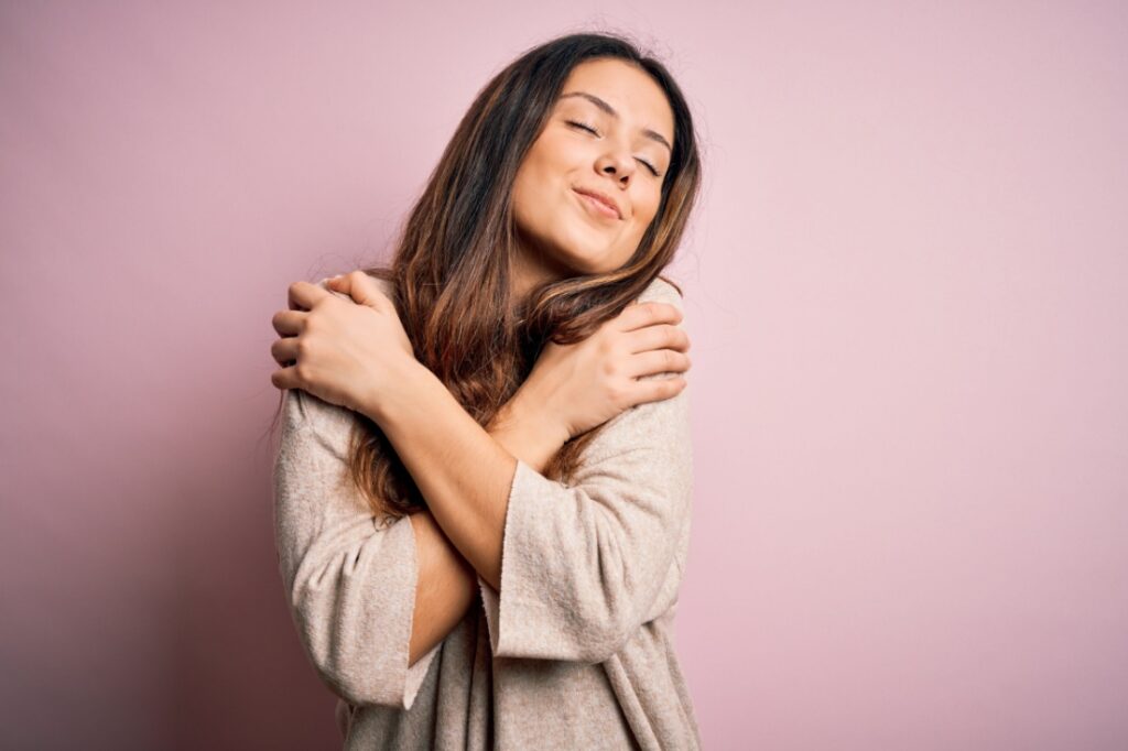 A woman hugging herself against a pink background.