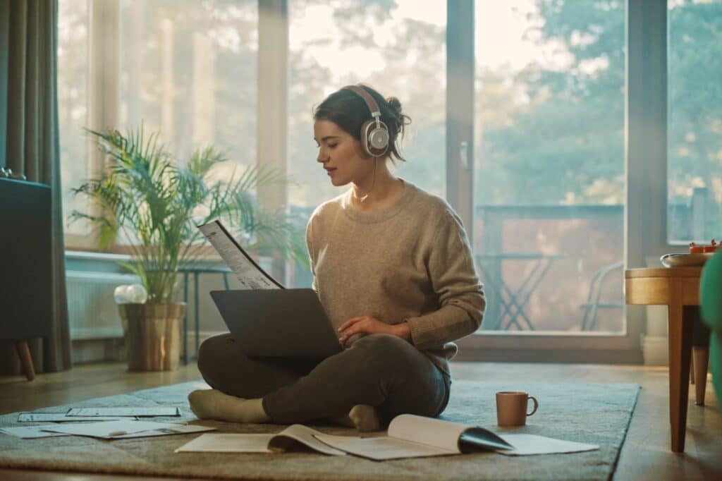 A woman on her living room floor, surrounded by books, looking at her laptop, wearing headphones.