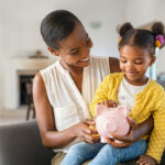 mother helping daughter sitting on lap putting money in piggy bank