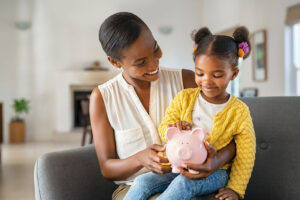 mother helping daughter sitting on lap putting money in piggy bank