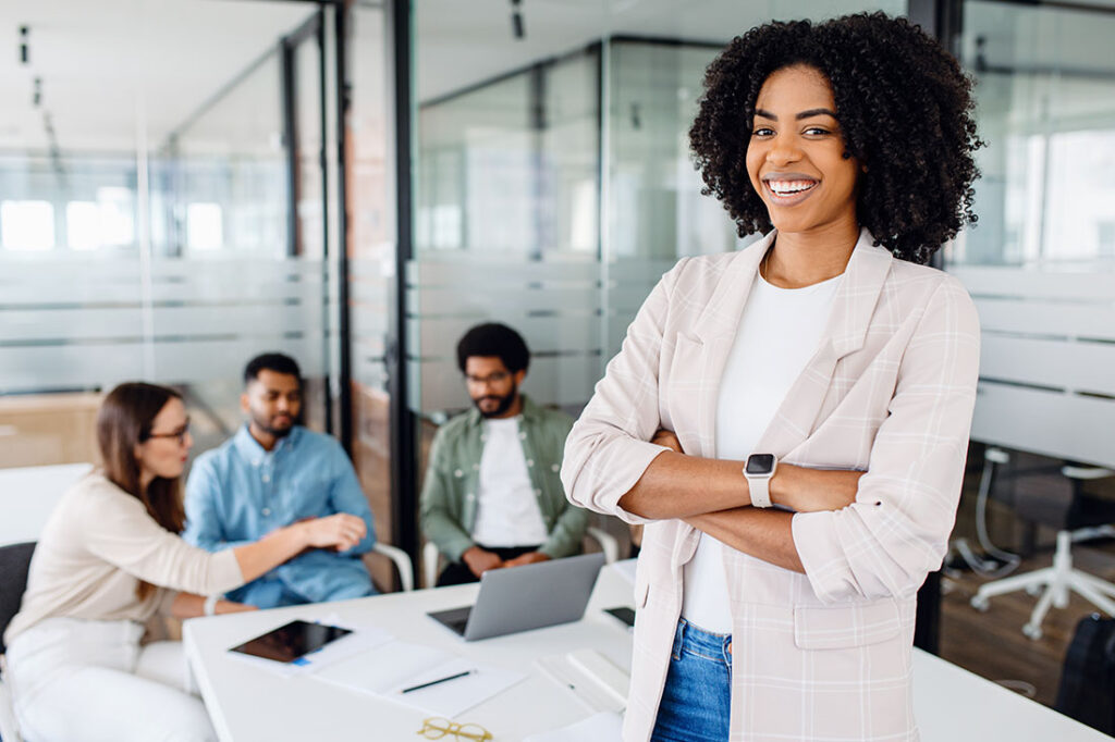 woman with a cheerful smile stands with arms crossed on the forefront in a modern office setting with her colleagues collaborating in the background,